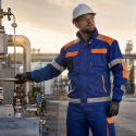 Industrieel medewerker in multinorm werkkleding met reflecterende strepen in een petrochemische raffinaderij tijdens golden hour, met leidingen en industriële installaties op de achtergrond.