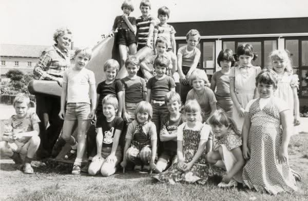 Klassenfoto uit 1980 van Evelyn, zes jaar oud, glimlachend tussen klasgenootjes. Aan de buitenkant een gewoon kind, vanbinnen al bezig zich aan te passen.