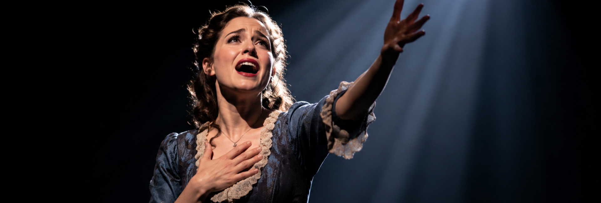 Emotionele musicalsolo in theaterlicht – VAYE Een musicalperformer zingt met uitgestrekte arm onder een sterke lichtbundel op een donkere scène.