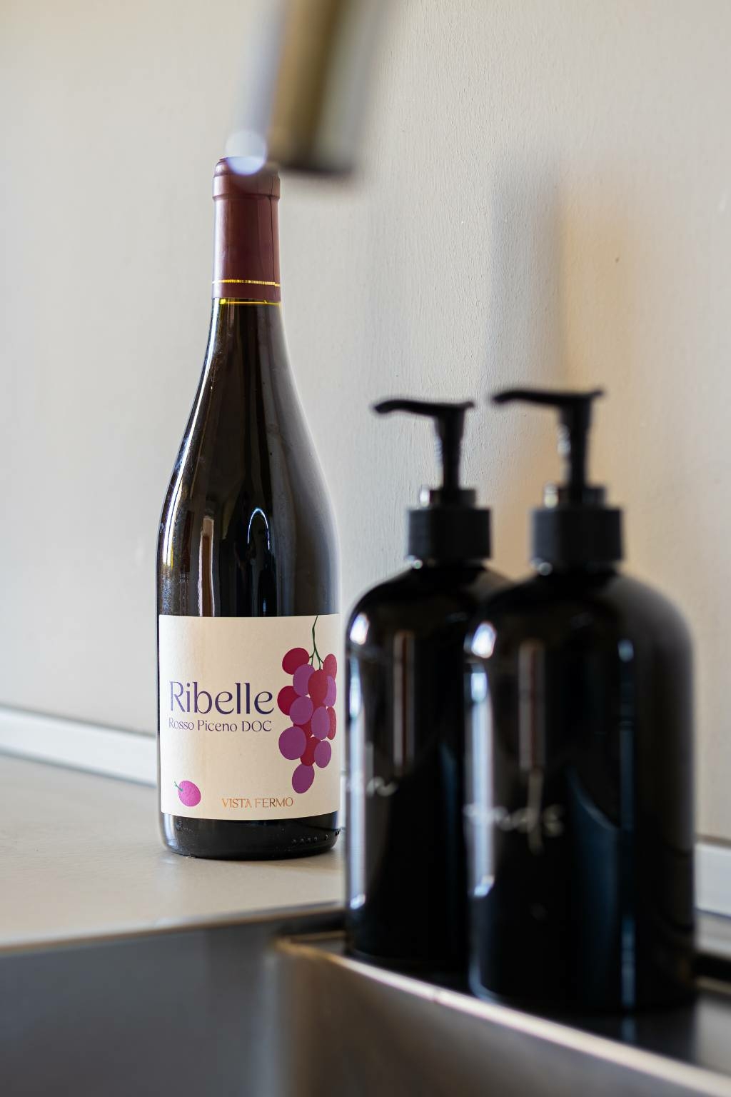 Fles eigen wijn Ribelle Rosso Piceno DOC van Vista Fermo op het aanrecht van Appartement Luce, naast verzorgingsproducten als welkom voor de gasten.