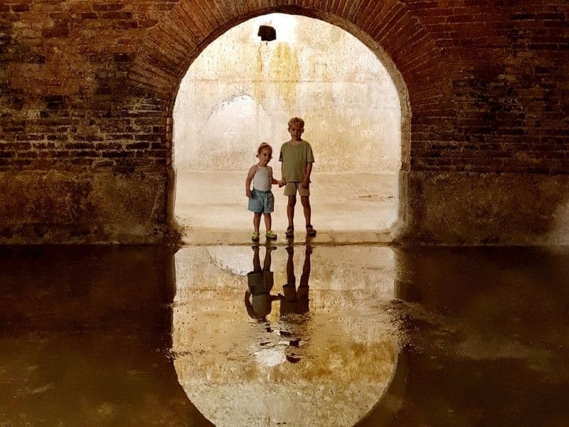 Twee kinderen staan in een oude stenen ruimte met een boog en weerspiegeling in het water tijdens een uitje in Fermo