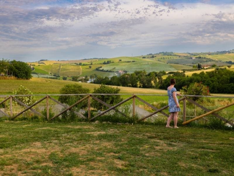Vrouw bij het houten hek van Vista Fermo geniet van het panorama over de groene heuvels en wijngaarden van Le Marche.