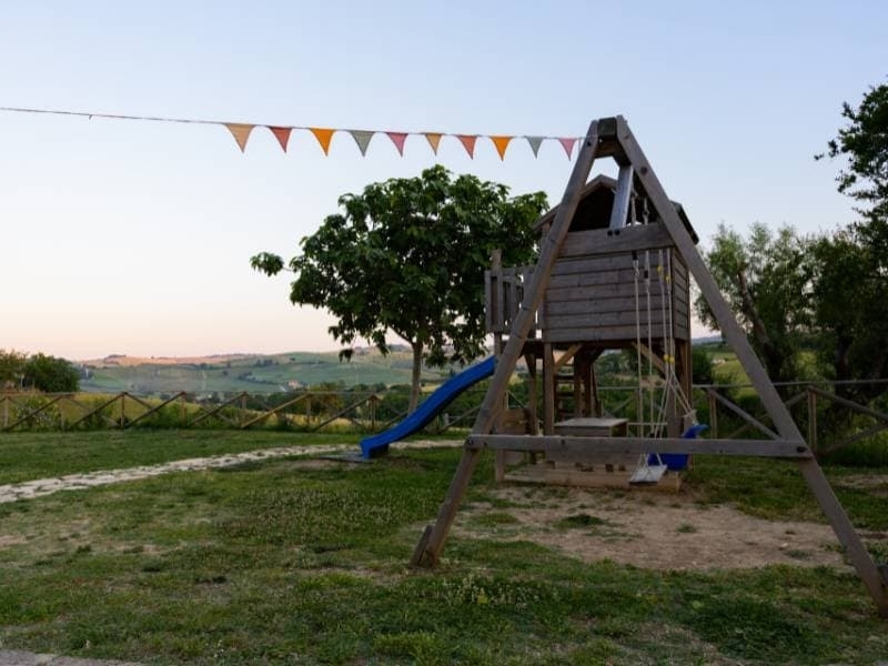 Houten speeltoestel met glijbaan en schommels op de kindvriendelijke speeltuin van Vista Fermo, met uitzicht over de heuvels van Le Marche bij avondlicht.