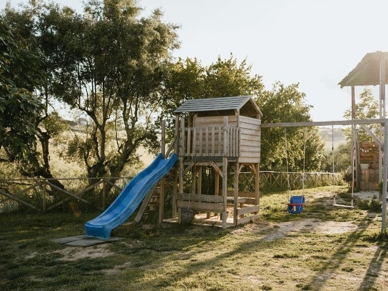 Houten speeltoestel met blauwe glijbaan en schommels op het terrein van Vista Fermo in de avondzon