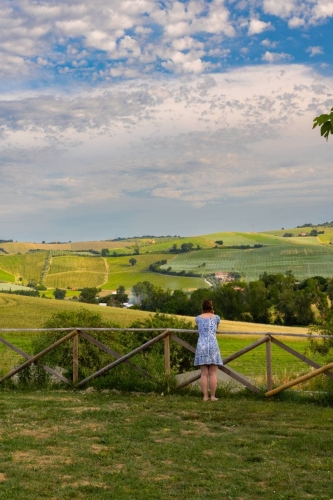 Vrouw kijkt vanaf Vista Fermo uit over de glooiende heuvels, wijngaarden en velden van Le Marche onder een wolkenlucht.