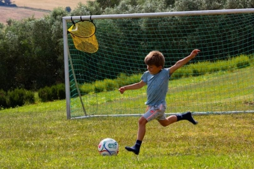 Kind schiet op doel op het voetbalveld van Vista Fermo, met olijfbomen en de heuvels van Le Marche op de achtergrond.