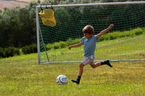 Kind schiet op doel op het voetbalveld van Vista Fermo, met olijfbomen en de heuvels van Le Marche op de achtergrond.