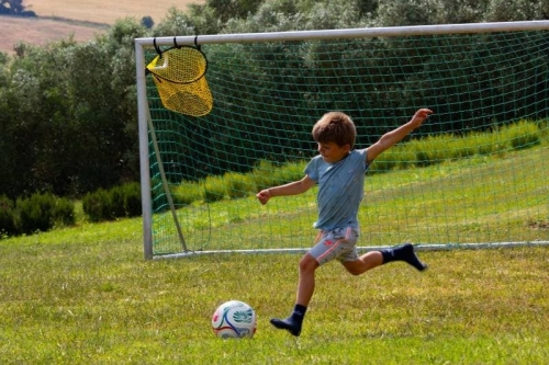 Kind schiet op doel op het voetbalveld van Vista Fermo, met olijfbomen en de heuvels van Le Marche op de achtergrond.