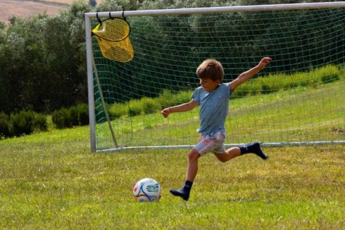 Kind schiet op doel op het voetbalveld van Vista Fermo, met olijfbomen en de heuvels van Le Marche op de achtergrond.