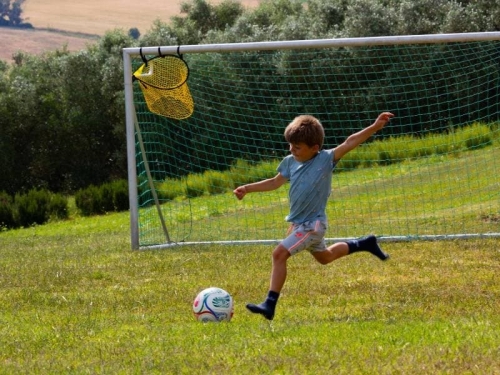 Kind schiet op doel op het voetbalveld van Vista Fermo, met olijfbomen en de heuvels van Le Marche op de achtergrond.