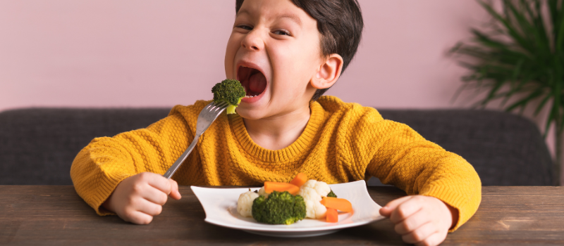 Een kind in een gele trui eet broccoli van een bord met groenten.