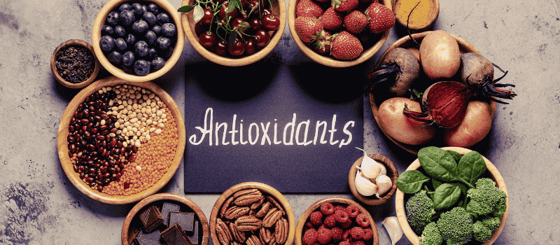 Diverse voedingsmiddelen rijk aan antioxidanten, zoals bessen, groenten, noten en kruiden, rond het woord 'Antioxidants'.