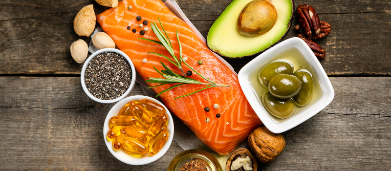 Verse zalm, avocado, olijven, noten en andere gezonde vetrijke voedingsmiddelen op een houten tafel.