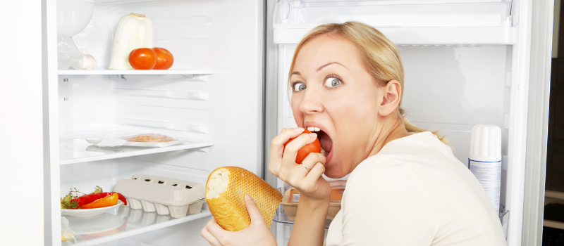 Vrouw eet een broodje en tomaat voor een open koelkast.