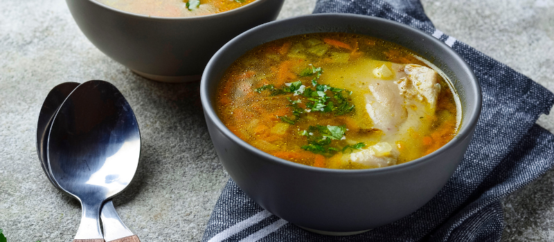 Een kom warme kippenbouillon met groenten en verse kruiden, geserveerd met een lepel.