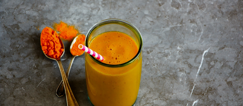 Glas met kurkuma-smoothie en twee lepels kurkumapoeder op een stenen ondergrond.