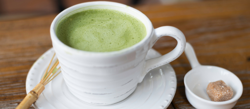 Kopje groene matcha-thee met een suikerklontje en kleine bamboe garde op een houten tafel.