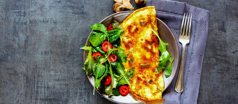 Gouden omelet met frisse salade van bladgroenten en rode tomaatjes op een grijs bord.