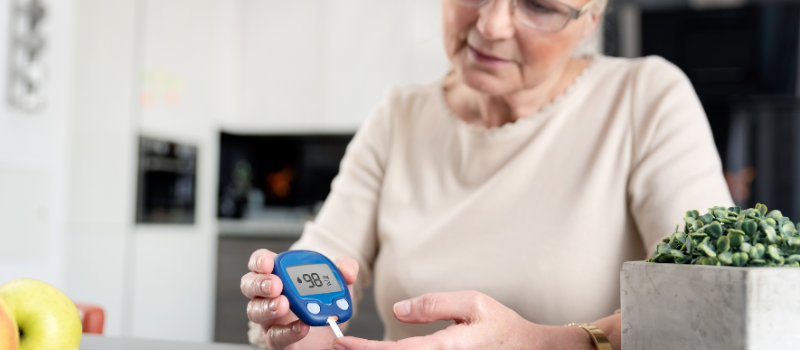 Oudere vrouw controleert bloedsuiker met een glucosemeter in een keuken.