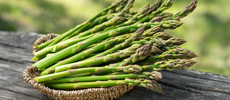 Een mand gevuld met verse groene asperges ligt op een houten tafel buiten.