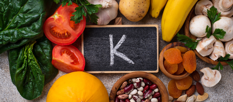 Diverse groenten en fruit rond krijtbord met “K”, rijke vitamine K bronnen