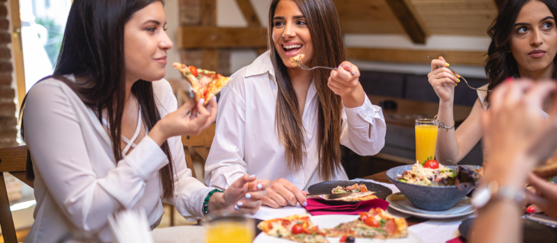 Groep vriendinnen geniet van pizza en salade aan een tafel met drankjes in een gezellige omgeving.