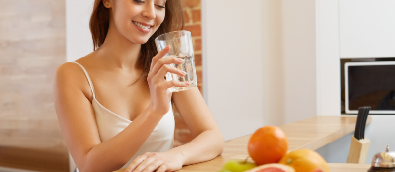 Vrouw drinkt glas water aan tafel met fruit, gezonde ochtendroutine