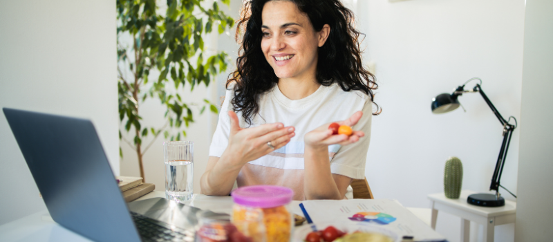 Vrouw eet een gezonde snack terwijl ze glimlacht en werkt achter een laptop.