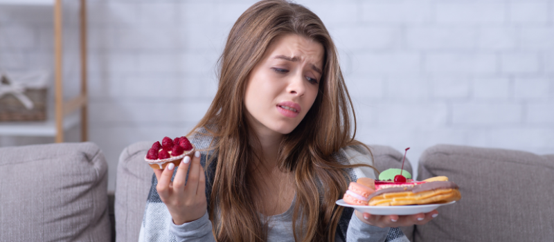 Vrouw kijkt gefrustreerd naar een bord met taart en fruit, twijfelend over haar keuze.