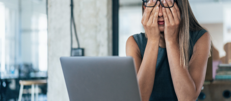 Vrouw met bril zit gestrest achter een laptop en houdt haar gezicht in haar handen.