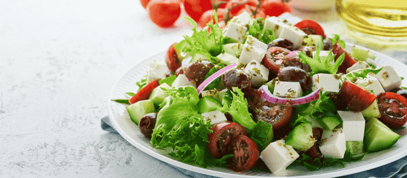 Een Griekse salade met feta, tomaten, komkommer, rode ui, olijven en groene sla op een bord.